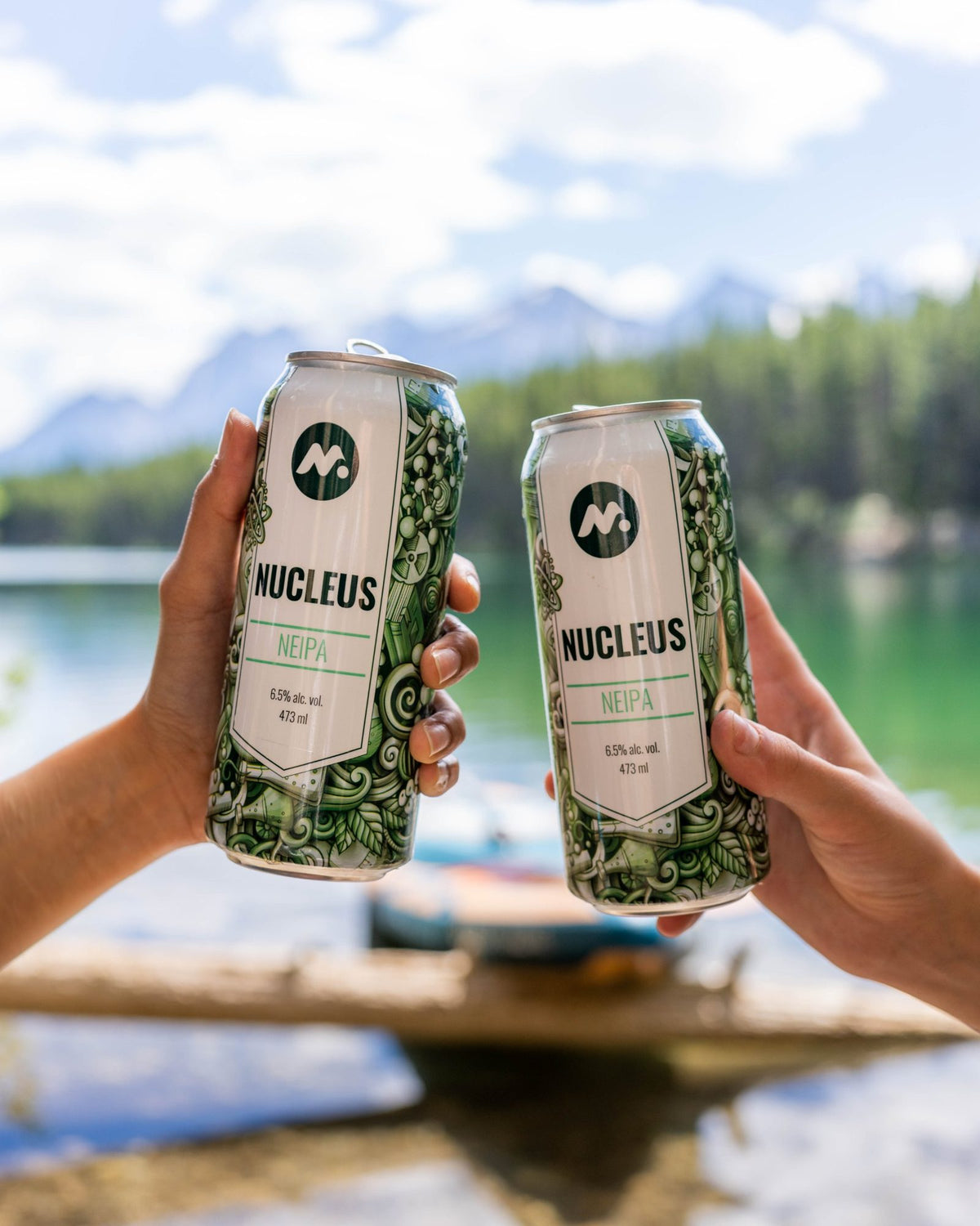 Two Nucleus NEIPA cans raised in a toast by a mountain lake